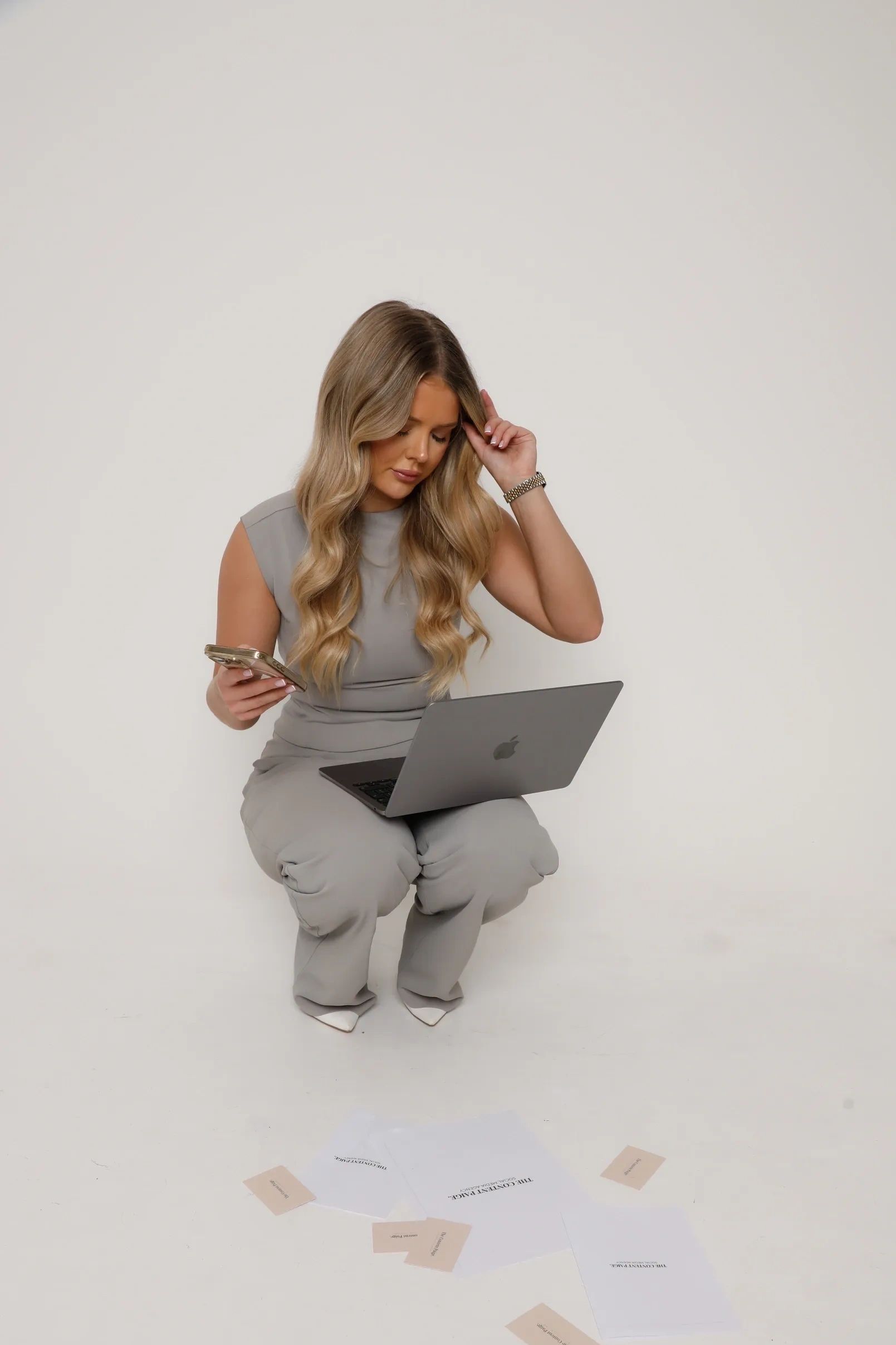 Woman in gray outfit multitasking with phone and laptop, surrounded by papers.