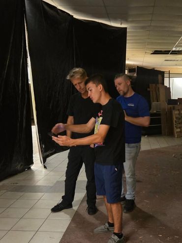 Three men looking at a phone indoors surrounded by black curtains.