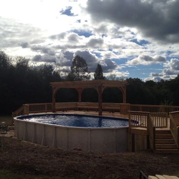 Above-ground pool with wooden deck and pergola under a cloudy sky.