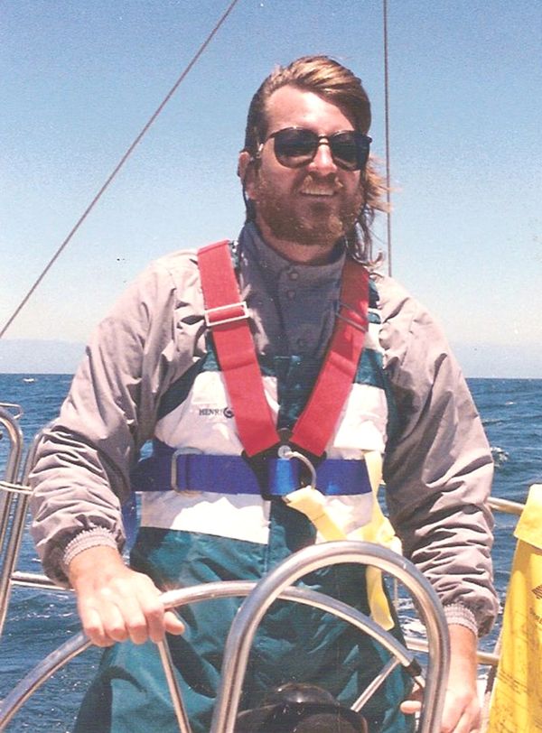 Clarke at the helm of a sailboat on the ocean, foul weather gear and lifejacket on, windswept