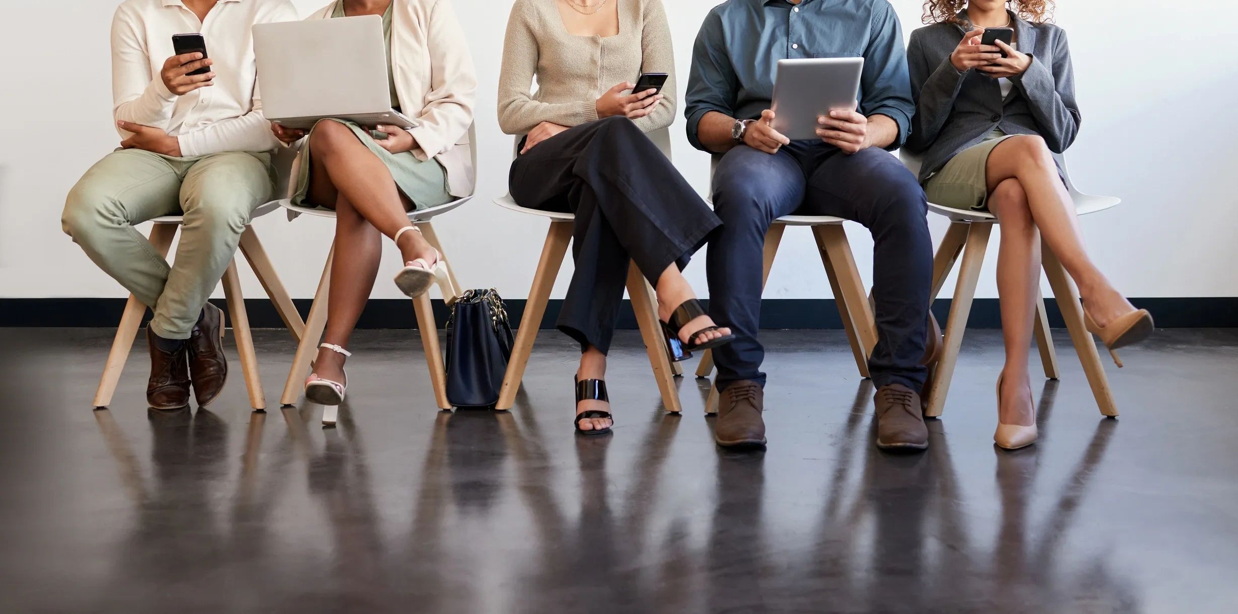 Five people sitting on chairs, using laptops, tablets, and phones in a waiting area.