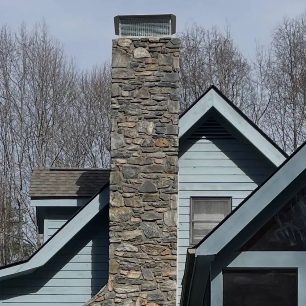 Stone chimney on blue house with leafless trees.