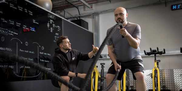 A trainer motivates a man working out with battle ropes in a gym.