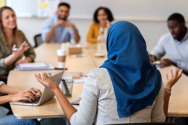 A woman in a blue hijab leads a meeting with diverse colleagues around a table.