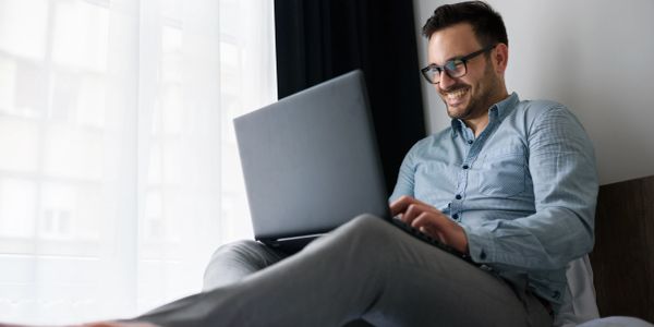 Man wearing glasses using a laptop and smiling while sitting on a bed.
