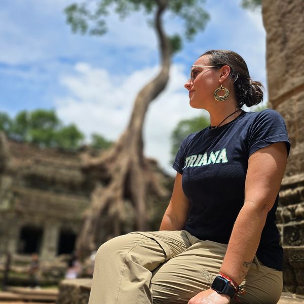 Woman sitting outdoors near ancient stone ruins with a large tree in the background.