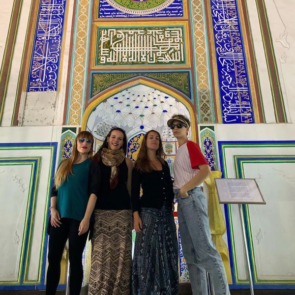 Four women posing inside a colorful, ornately decorated building with Arabic calligraphy.
