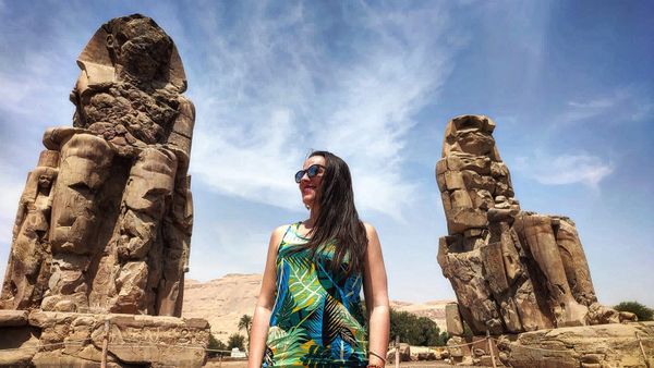 Woman in colorful dress standing between two ancient stone statues under a blue sky.