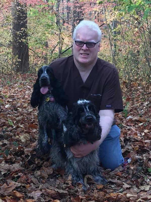 Man with glasses kneeling in autumn leaves holding two black and gray dogs.