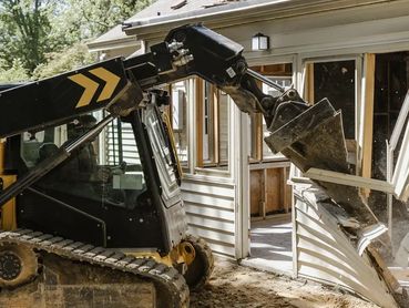 A yellow excavator demolishes a white house wall with a window.
