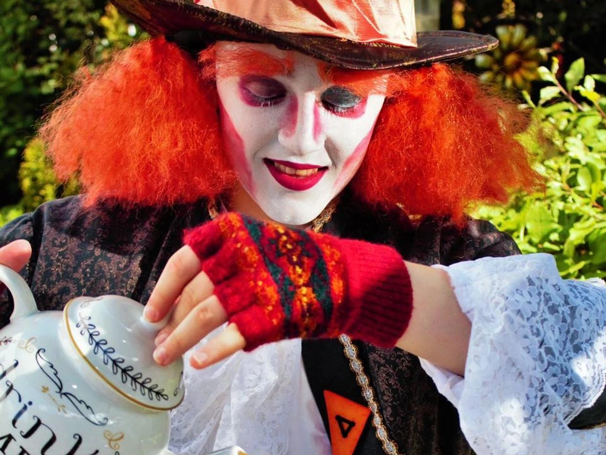 Person dressed as a colorful Mad Hatter pouring tea outdoors.