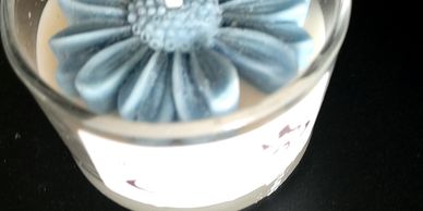 A candle in a glass container with a blue flower-shaped wax decoration on top.