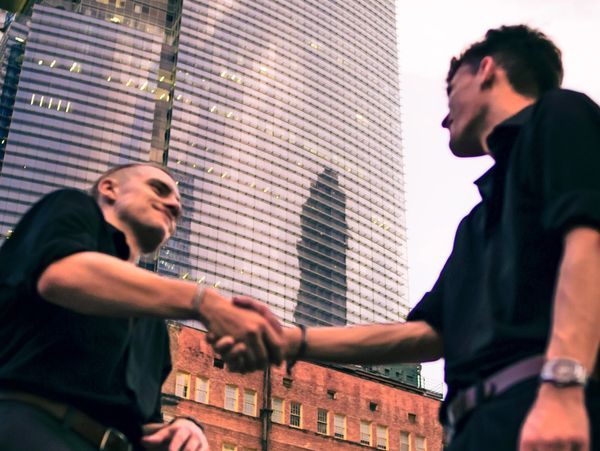two men meet and shake hands with each other, with a building in the background.