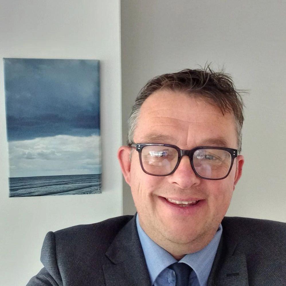 Smiling man in glasses and suit with ocean artwork behind him.