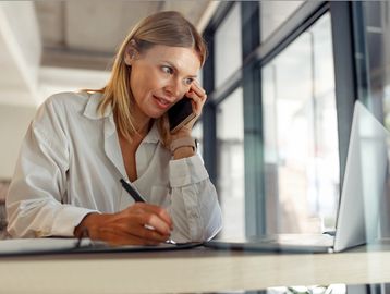 Woman talking on phone and writing notes at her laptop.