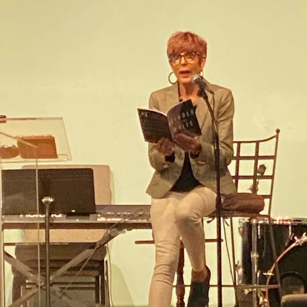 Person reading from a book on stage with microphone and musical instruments nearby.