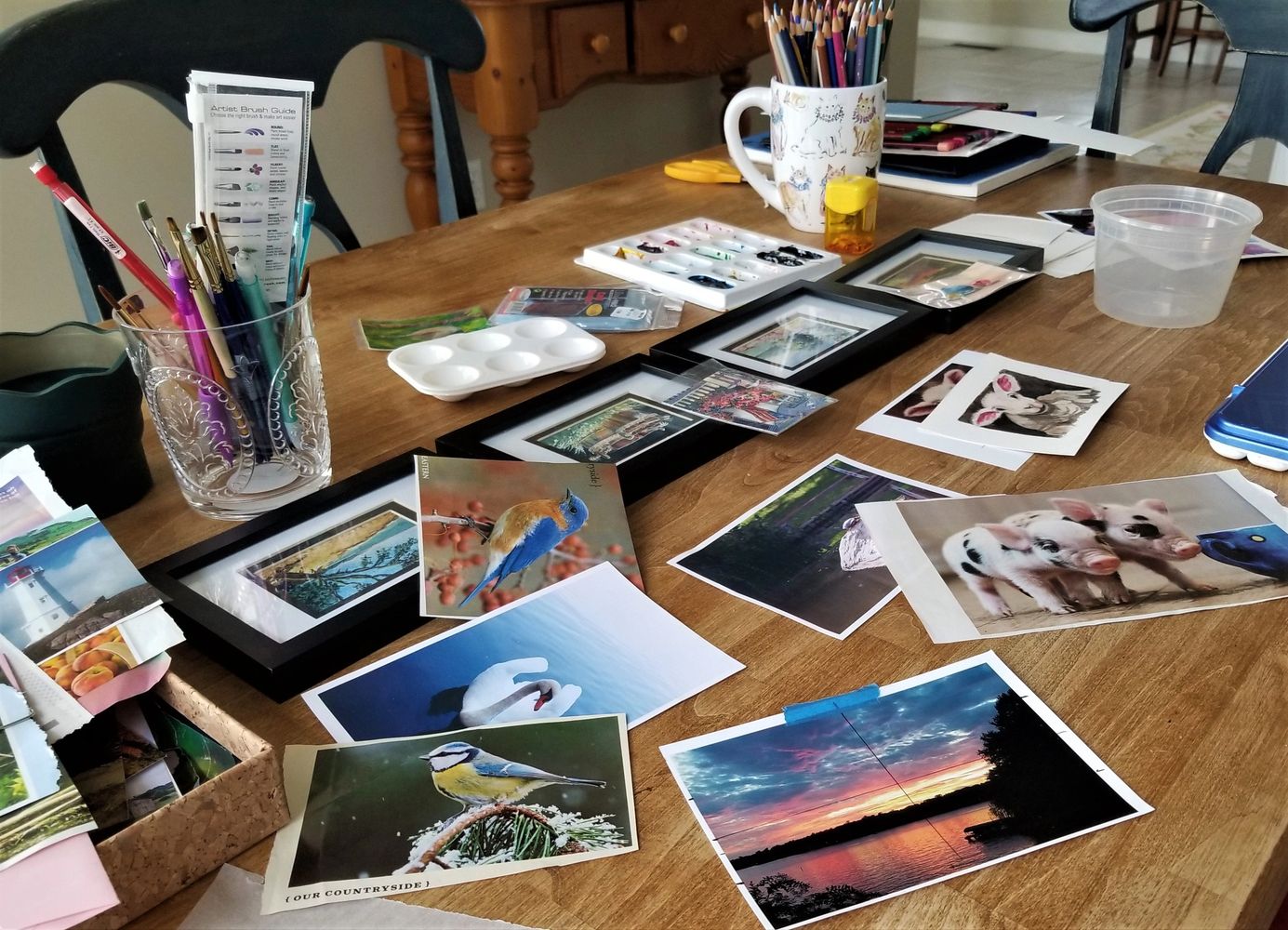 A kitchen table filled with inspiration photographs, painted pictures, pencils, brushes and paint