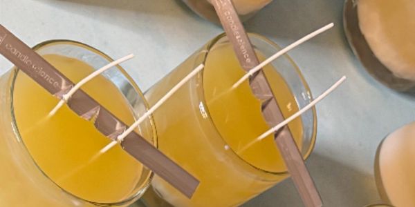 Glass containers filled with yellow wax and wooden wick holders for candle making.