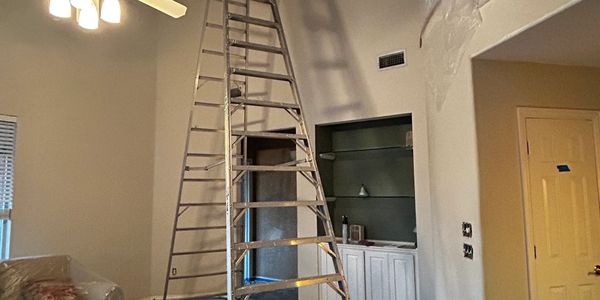A tall ladder extends inside a room with beige walls and ceiling fan.