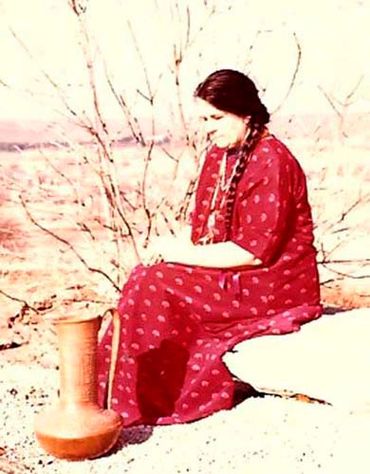 Inez Running-rabbit wearing a red dress, sitting with a pot she made.