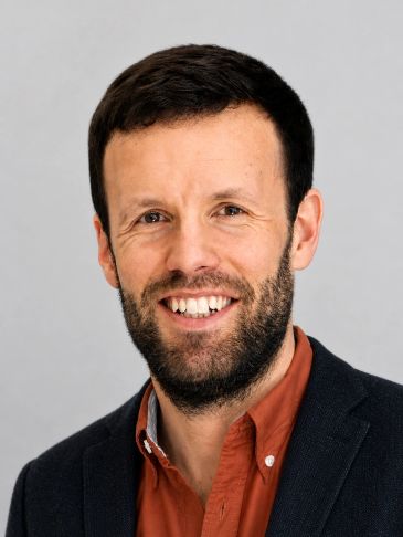 Smiling man with beard in casual shirt and blazer against a plain background.