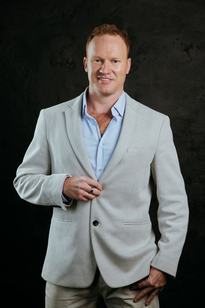 Confident man in a light grey blazer smiling against a dark background.