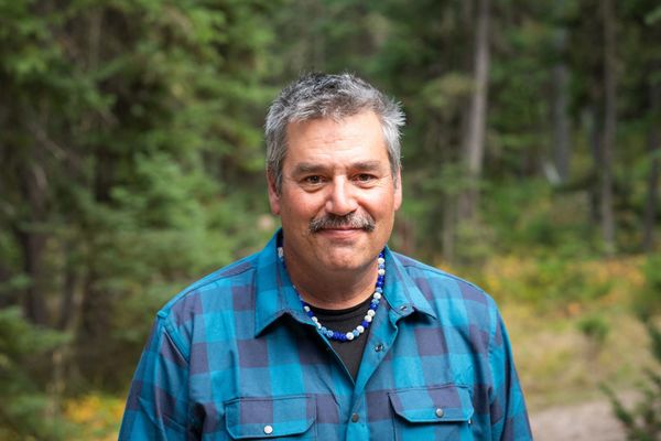 Man smiling outdoors in a blue checkered shirt and beaded necklace.
