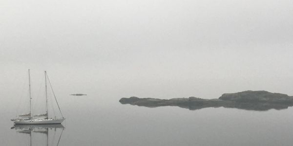 A solitary sailboat on calm water near a small island in fog.