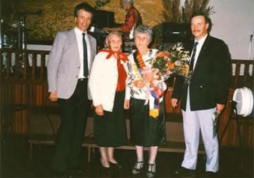 Four people posing indoors, two women holding flowers and wearing sashes.