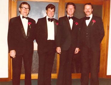 Four men in tuxedos with red boutonnieres posing indoors in front of a wooden door.
