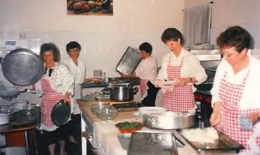 Five women in aprons working together in a kitchen preparing food.