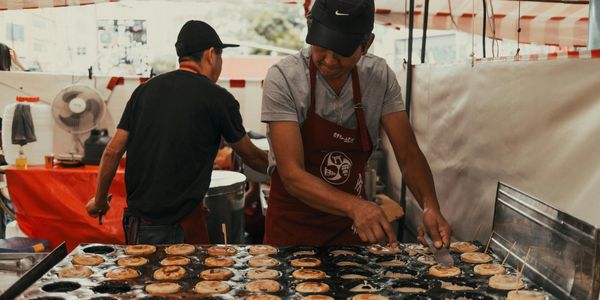 Two men cooking round pastries on a large griddle at a food stall.