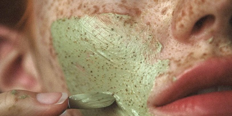 Close-up of a person applying a green facial mask with tiny exfoliating particles.