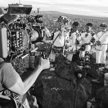 Filming indigenous ritual with traditional attire and a stone sculpture outdoors.