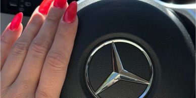 Hand with red nails near Mercedes-Benz steering wheel logo.
