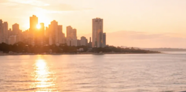 Sunset over city skyline reflected on calm water from a waterfront railing.
