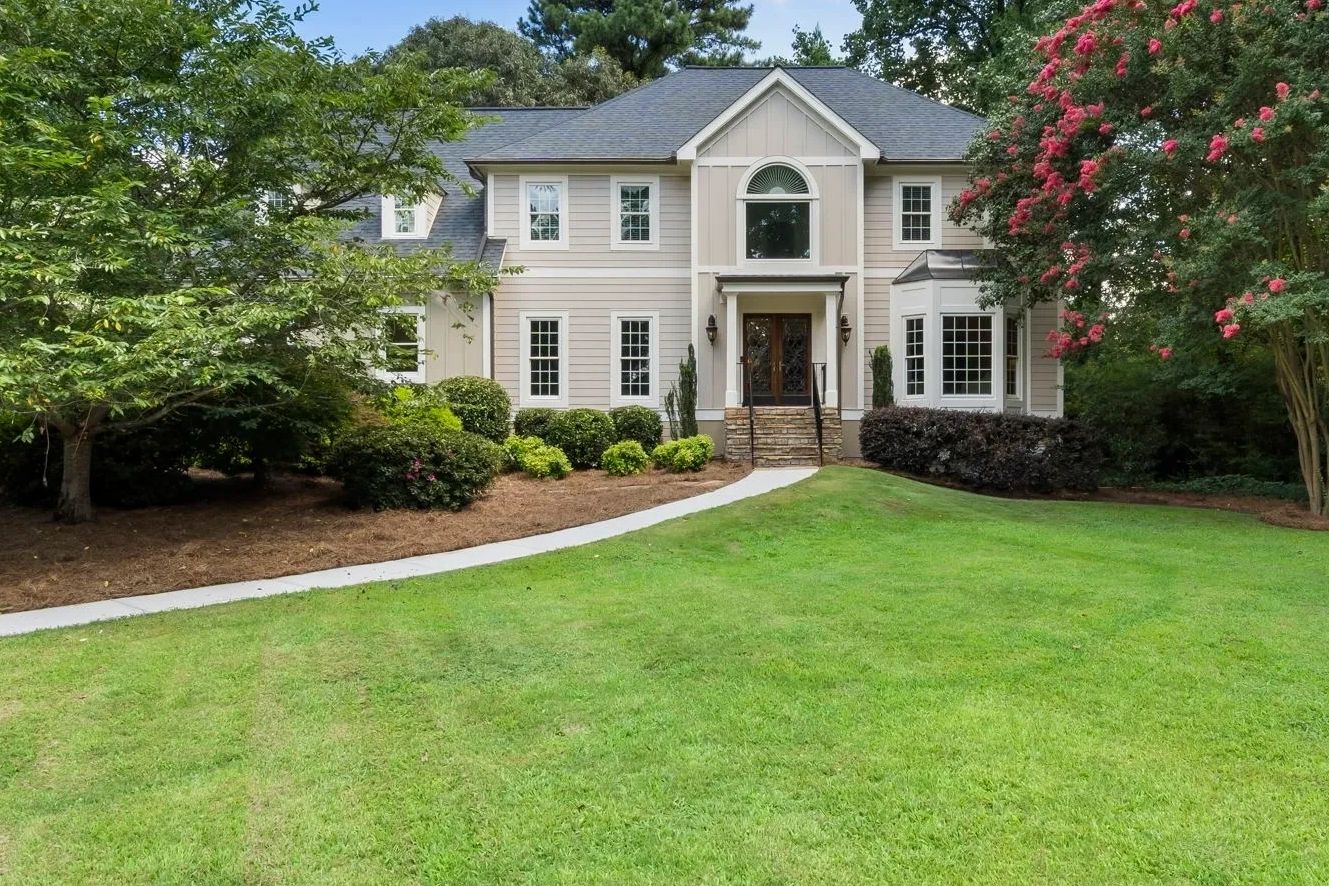 Two-story beige house with manicured lawn and trees.