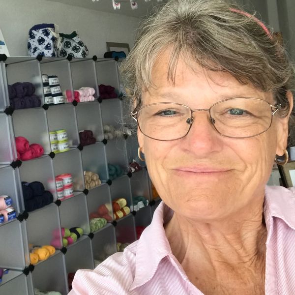 Smiling woman with glasses in front of colorful yarn shelves.