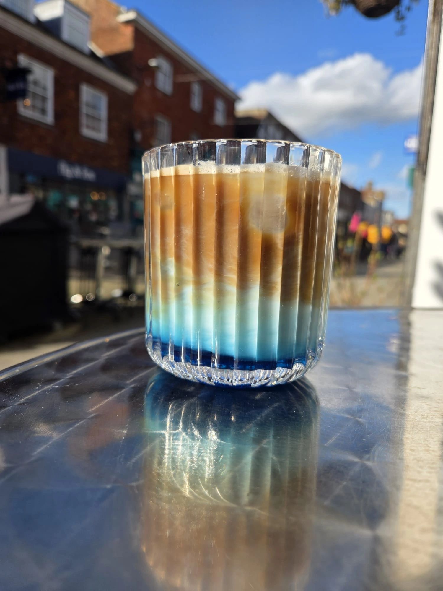 Iced coffee in a clear glass with blue hues at the bottom on a reflective table.