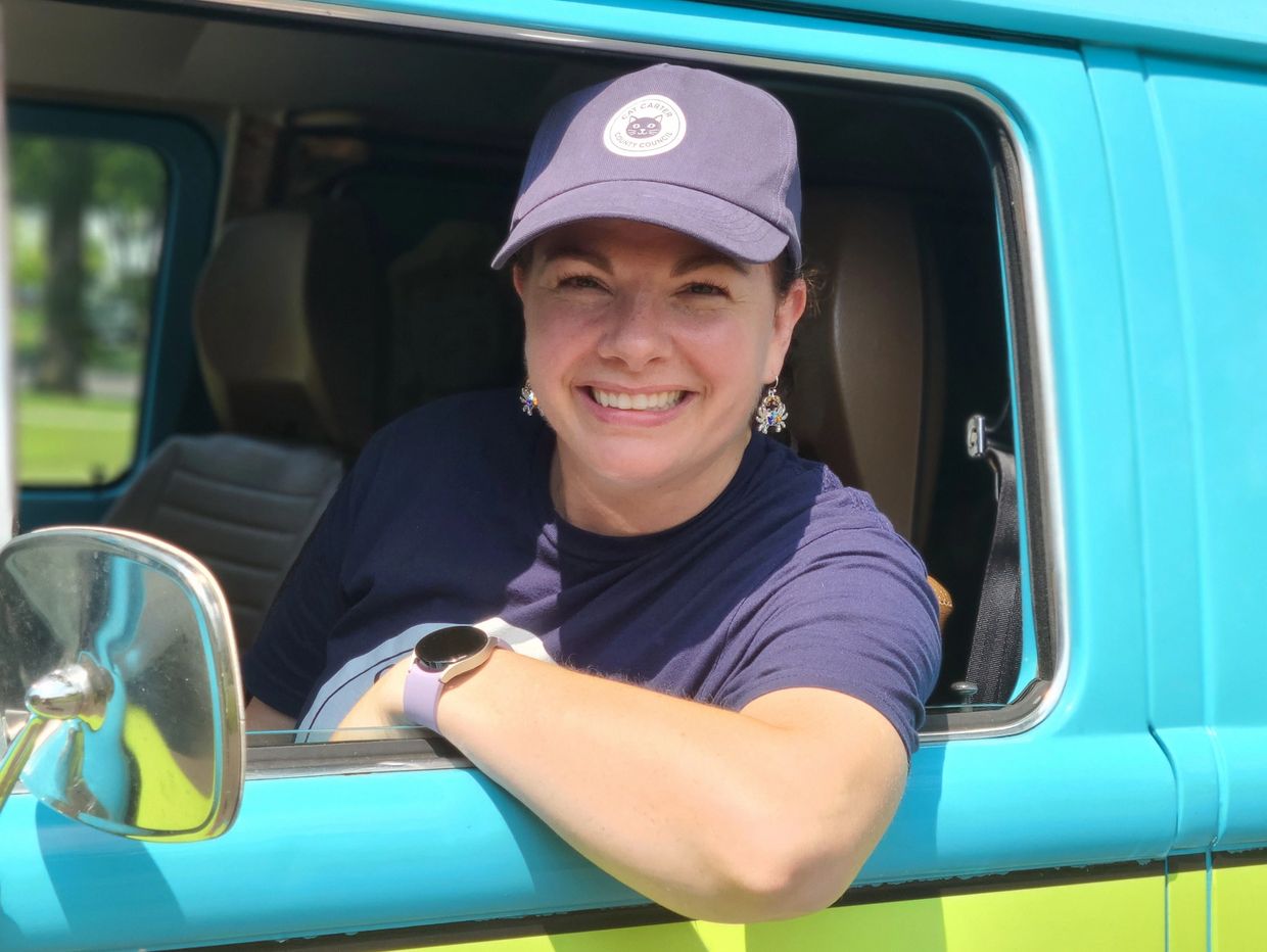 Smiling woman wearing a cap leans out of a blue vehicle window.