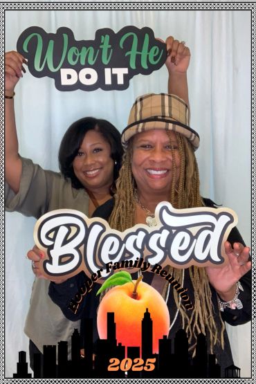 Two women smiling and holding signs saying 'Won't He Do It' and 'Blessed' at a family reunion.