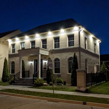 Illuminated two-story brick house at night with outdoor lighting and greenery.