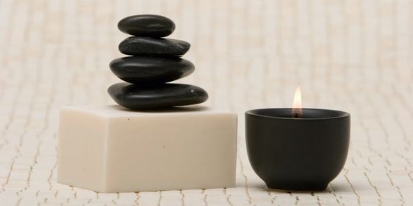 Stacked black stones on a white block next to a lit black candle in a serene setting.