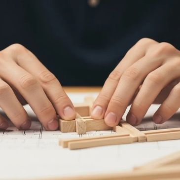 A student's hands carefully assemble a small wooden architectural model over blueprints, symbolizing the precision and skill of the trades.