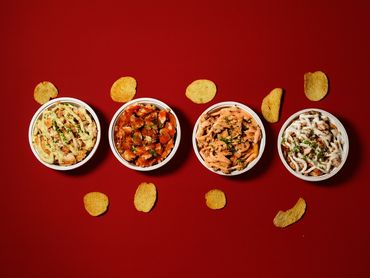 Four bowls of seasoned chicken with different sauces, surrounded by potato chips on a red background.