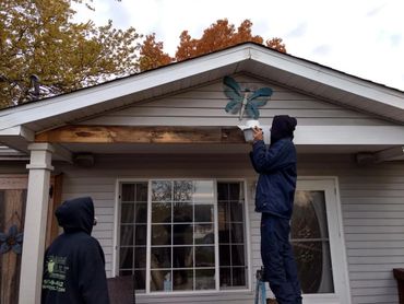 Two people working on a porch roof, one on a ladder painting a butterfly decoration.