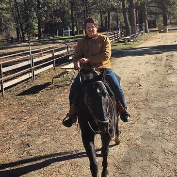 Person riding a black horse on a dirt path surrounded by trees and fences.