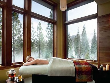 Woman relaxing on massage table in a serene room with large windows overlooking pine trees.