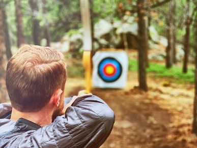 Man aiming a bow at a colorful archery target in the woods.