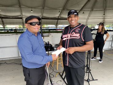 Ollie in a blue, long sleeve button down shirt and sunglasses and hat, holds a check with another ma